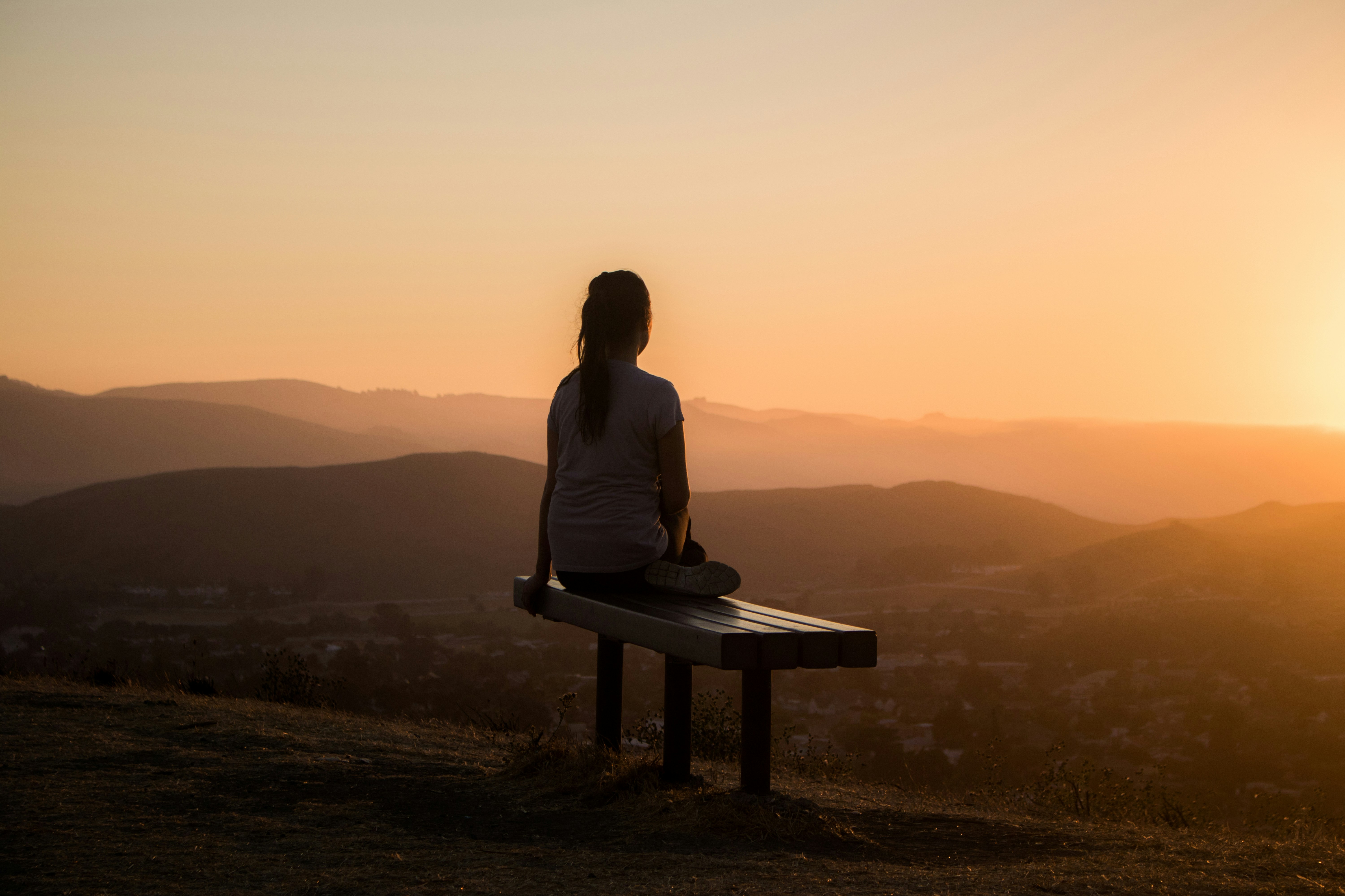 Person sitzt allein auf einer Holzbank auf einem Hügel und blickt in den Sonnenuntergang über einer hügeligen Landschaft